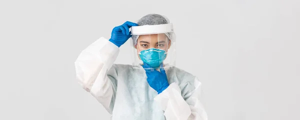 Covid-19, coronavirus disease, healthcare workers concept. Confident serious-looking female asian doctor, put on face shield and respirator, personal protective equipment, white background — Stock Photo, Image