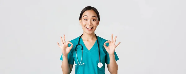 Healthcare workers, preventing virus, quarantine campaign concept. Excited and pleased asian female doctor, nurse in scrubs with stethoscope, showing okay gesture, white background — Stock Photo, Image