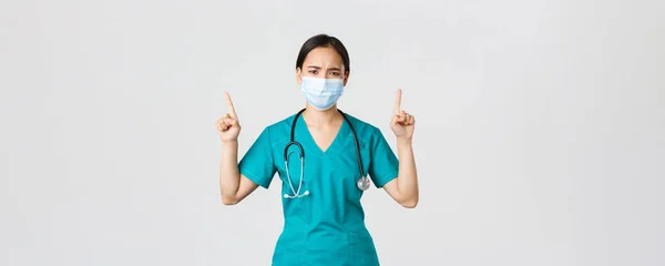 Covid-19, maladie du coronavirus, concept des travailleurs de la santé. Sombre déçu infirmière asiatique, médecin dans le masque médical et les gommages, les doigts pointant vers le haut et fronçant les sourcils perplexe, l'air frustré — Photo