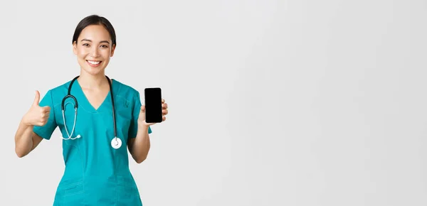 Covid-19, trabajadores de la salud y concepto de medicina en línea. Retrato de joven asiática médico, médico en matorrales mostrar pulgares hacia arriba y la pantalla del teléfono inteligente, promover la aplicación, fondo blanco — Foto de Stock