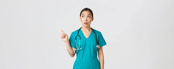 Covid-19, sağlık çalışanları, salgın konsepti. Şaşırmış ve şaşırmış kadın Asyalı doktor sol üst köşeyi işaret ediyor. Şaşkın Koreli doktor reklamla pankarta bakıyor — Stok fotoğraf