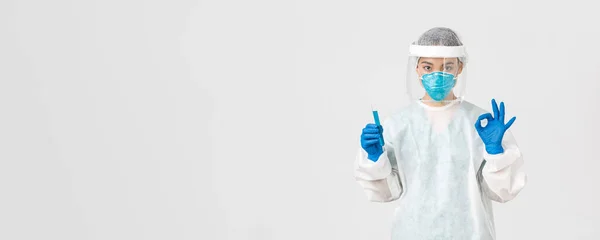 Covid-19, coronavirus disease, healthcare workers concept. Serious asian female doctor, professional researcher in personal protective equipment showing okay gesture, holding test-tube with vaccine — Stock Photo, Image