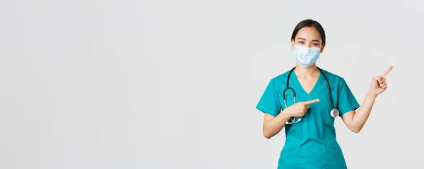 Covid-19, coronavirus disease, healthcare workers concept.快乐的亚洲女医生，戴着洗涤器和面罩的医护人员，手指指向右上角，展示广告 — 图库照片