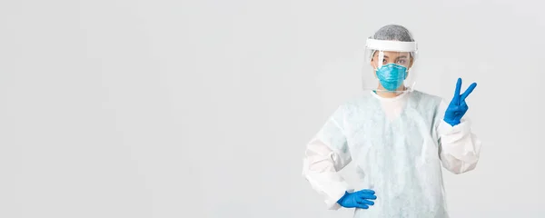 Covid-19, coronavirus ziekte, gezondheidswerkers concept. Sassy aziatische vrouwelijke arts, arts of tech lab in persoonlijke beschermingsmiddelen tonen vrede teken, staande witte achtergrond — Stockfoto