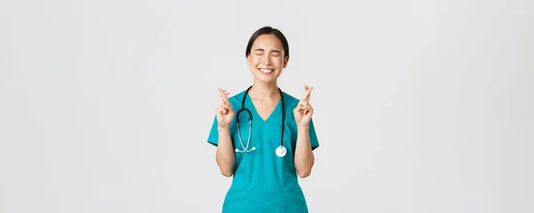 Covid-19, egészségügyi dolgozók, világjárvány. Optimista boldog és reményteljes ázsiai női ápolónő, gyakornok várja az előléptetést, keresztujjakkal sok szerencsét és csukott szemmel, mosolyogva izgatott — Stock Fotó