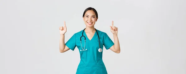 Covid-19, healthcare workers, pandemic concept. Smiling beautiful asian female nurse, doctor in scrubs pointing and looking up with satisfied grin, happy to show awesome promo offer, white background — Stock Photo, Image