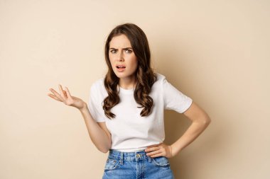 Confused woman looking clueless and annoyed, cant understand, asking whats problem, standing over beige background