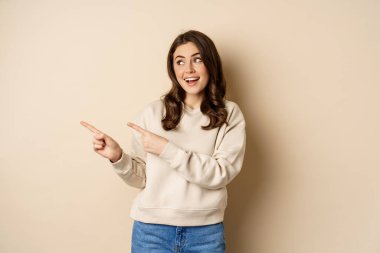 Cheerful attractive female model pointing fingers left, showing store advertisement, banner or logo, standing in sweater over beige background