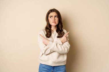Complicated and indecisive woman pointing sideways, cant make choice, struggle with decision, choosing between two options, standing over beige background