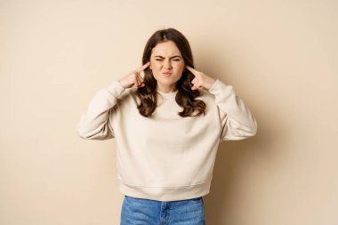 Annoyed and angry woman shut ears, complaining at loud noisy music noise, standing over beige background