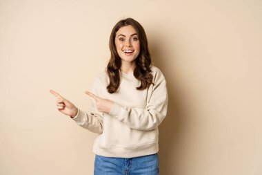 Cheerful attractive female model pointing fingers left, showing store advertisement, banner or logo, standing in sweater over beige background