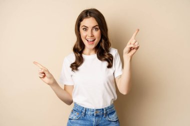 Happy authentic girl smiling, pointing fingers sideways, showing left and right banner, demonstrating promo, standing against beige background