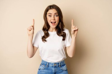 Enthusiastic young woman, female customer pointing fingers up and smiling, showing banner or logo, standing against beige background