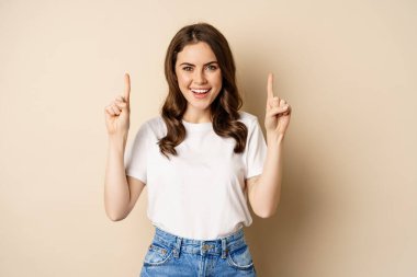 Enthusiastic young woman, female customer pointing fingers up and smiling, showing banner or logo, standing against beige background