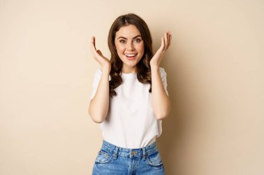 Surprise. Happy young woman open eyes with hands and waiting for gift, standing over beige background