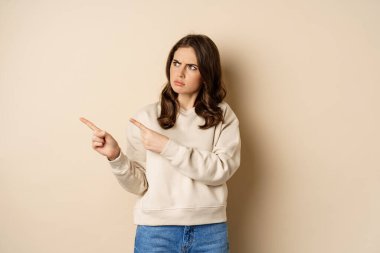 Woman pointing left and staring confused at banner copy space, strange logo, standing over beige background