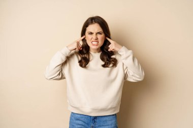 Annoyed and angry woman shut ears, complaining at loud noisy music noise, standing over beige background