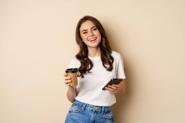 Smiling woman using mobile phone app and credit card, concept of online shopping, contactless payment and delivery, standing over beige background