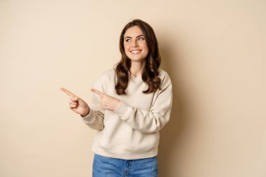 Beautiful girl pointing and looking left, laughing and smiling happy while showing advertisement, logo or store banner, beige background