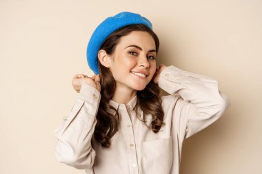 Stylish modern girl put on trendy hat on head and smiling, going out, posing against beige background