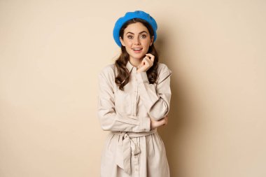 Image of stylsh beautiful woman looking surprised, shocked reaction at camera, posing in trench coat against beige background