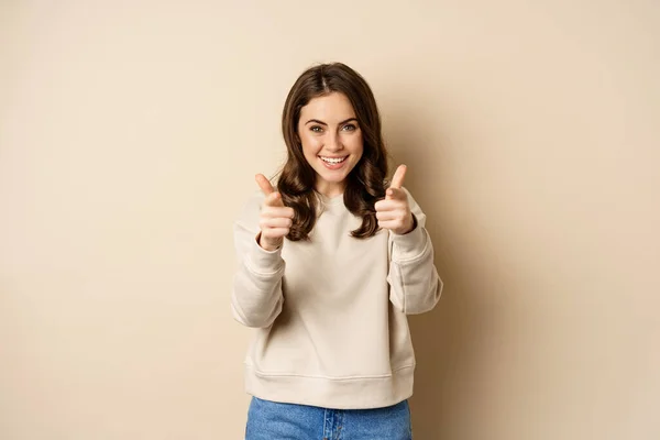 Smiling happy woman choosing you, pointing fingers at camera and laughing, inviting people, standing over beige background — 스톡 사진