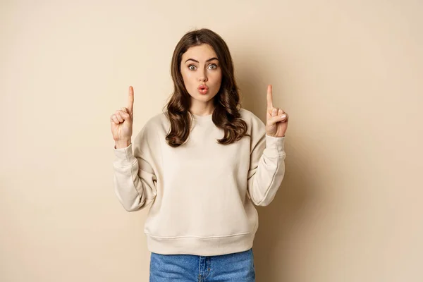 Surprised beautiful girl showing advertisement, pointing fingers on top, store sale or website link up, standing over beige background — Foto de Stock