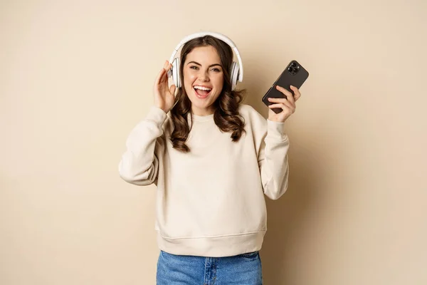 Happy caucasian woman listening music in headphones on smartphone app, dancing and having fun, standing over beige background — Fotografia de Stock