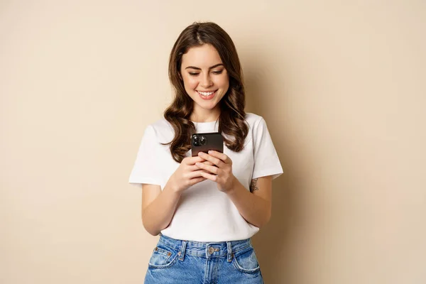 Ung modern flicka chatta på app, med hjälp av smartphone app och leende, står över beige bakgrund — Stockfoto