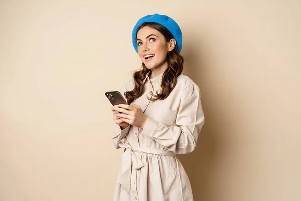 Menina moderna usando smartphone, sorrindo e posando feliz com telefone celular, de pé sobre fundo bege — Fotografia de Stock