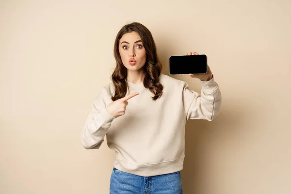 Imagem da menina apontando o dedo, mostrando tela horizontal do telefone móvel e olhando espantado com o aplicativo, de pé sobre fundo bege — Fotografia de Stock
