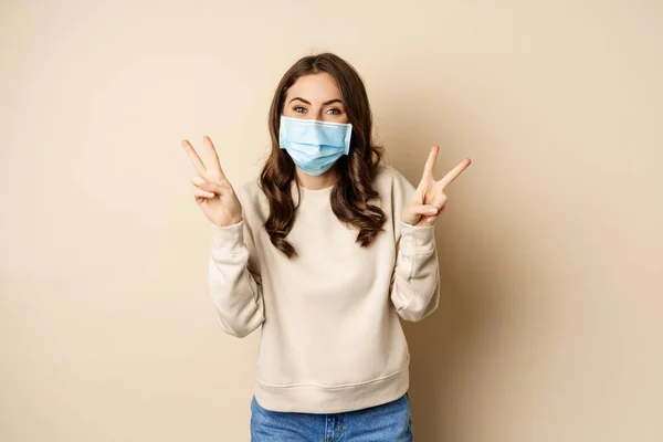 Covid-19, pandemic and quarantine concept. Beautiful adult woman in face medical mask, showing peace, v-sign gesture, standing over beige background — Φωτογραφία Αρχείου