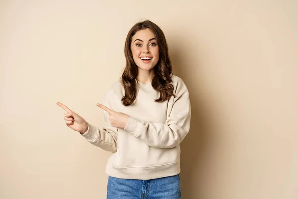 Modelo femenino atractivo alegre que señala los dedos a la izquierda, mostrando anuncio de la tienda, bandera o logotipo, de pie en suéter sobre fondo beige — Foto de Stock