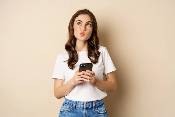 Retrato de mulher pensando, segurando telefone celular e olhando para cima com o rosto pensativo, de pé sobre fundo bege — Fotografia de Stock