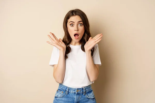 Portrait of stylish young woman gasping, looking surprised and amazed, impressed by smth, standing in t-shirt and jeans over beige background — Stock fotografie