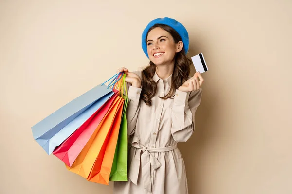 Portrat of trendy feminine girl posing with shopping bags from store and credit card, paying contactless, buying with discount on sale, beige background — Stockfoto