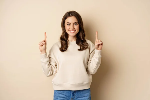 Ενθουσιασμένο κορίτσι που δείχνει τα δάχτυλα προς τα πάνω, δείχνει την ανακοίνωση, promo κείμενο ή το λογότυπο προς τα πάνω, στέκεται πάνω από το μπεζ φόντο — Φωτογραφία Αρχείου