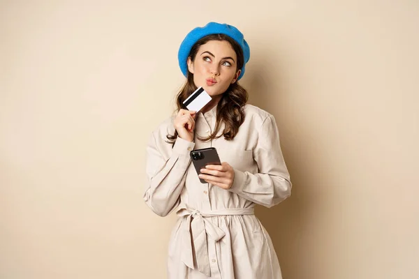 Портрет модной женщины в пальто, держащей смартфон, показывающей кредитку, делающей покупки онлайн, заказывающей что-то на мобильном телефоне, стоящей на бежевом фоне — стоковое фото