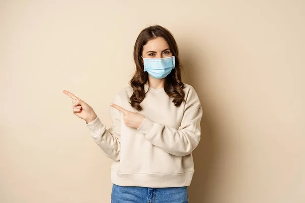 Health and covid-19 concept. Cute brunette woman in medical face mask, pointing fingers left at logo, banner, showing copy space, standing over beige background — ストック写真