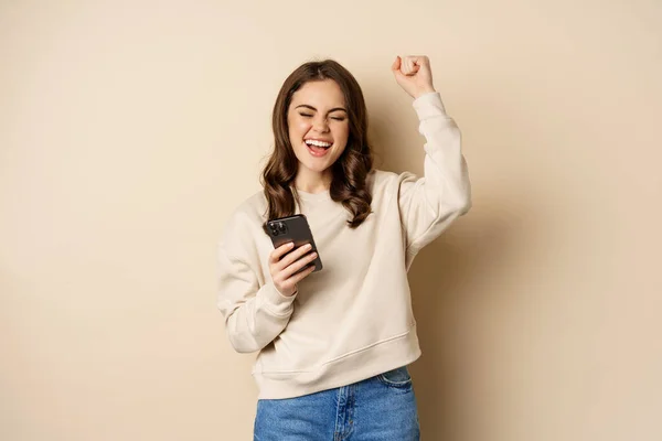 Glad brunett flicka som håller smartphone och jublar, vinna, fira vinna på mobiltelefon app, står över beige bakgrund — Stockfoto