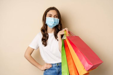 Covid-19 pandemic and lifestyle concept. Young woman posing in medical face mask with shopping bags from mall, vaccinated girl going in store in personal protective equipment, beige background
