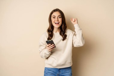 Akıllı telefon tutan, tezahürat yapan, kazanan, kutlayan mutlu esmer kız cep telefonu uygulamasında bej arka planda duruyor.