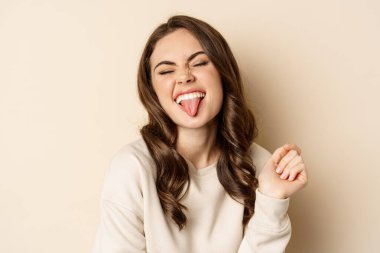 Close up of carefree happy woman, showing tongue and having fun, posing in sweater over beige background
