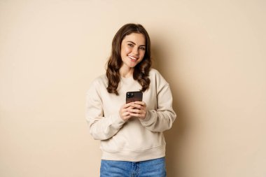 İnsanlar ve hücresel teknoloji. Güzel stil sahibi bir kadın cep telefonu kullanıyor, akıllı telefon uygulaması kullanıyor, gülümsüyor ve ekrana bakıyor, bej arka planda duruyor.