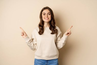 Cheerful caucasian woman showing directions, two ways, pointing sideways at variants, choices for customer, standing over beige background
