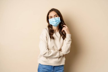 Covid-19, pandemic and quarantine concept. Beautiful modern woman in medical face mask, cross arms on chest, looking confident, beige background