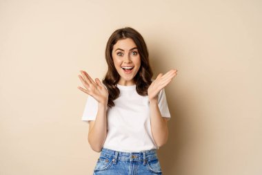 Portrait of happy young woman rejoicing, clapping hands and smiling, applause, standing over beige background