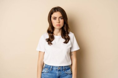 Sad and tired brunette woman looking drained and unamused, standing with pokerface against beige background