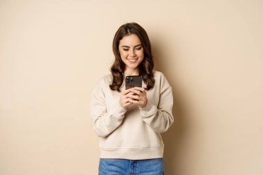 People and cellular technology. Beautiful stylish woman using mobile phone, smartphone app, smiling and looking at screen, standing over beige background