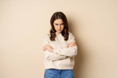 Angry girlfriend sulking, cross arms on chest and look offended, staring moody at camera, standing over beige background